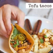 Text overlay: Tofu tacos. Hand holding tofu taco on plate with vegan breakfast sausage links and potato hash.