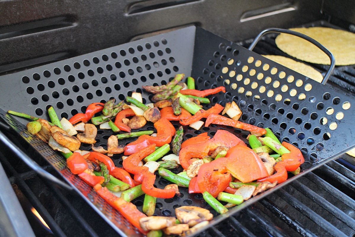 Asparagus, mushrooms, onions, and bell peppers in grill basket by corn tortillas on grate.