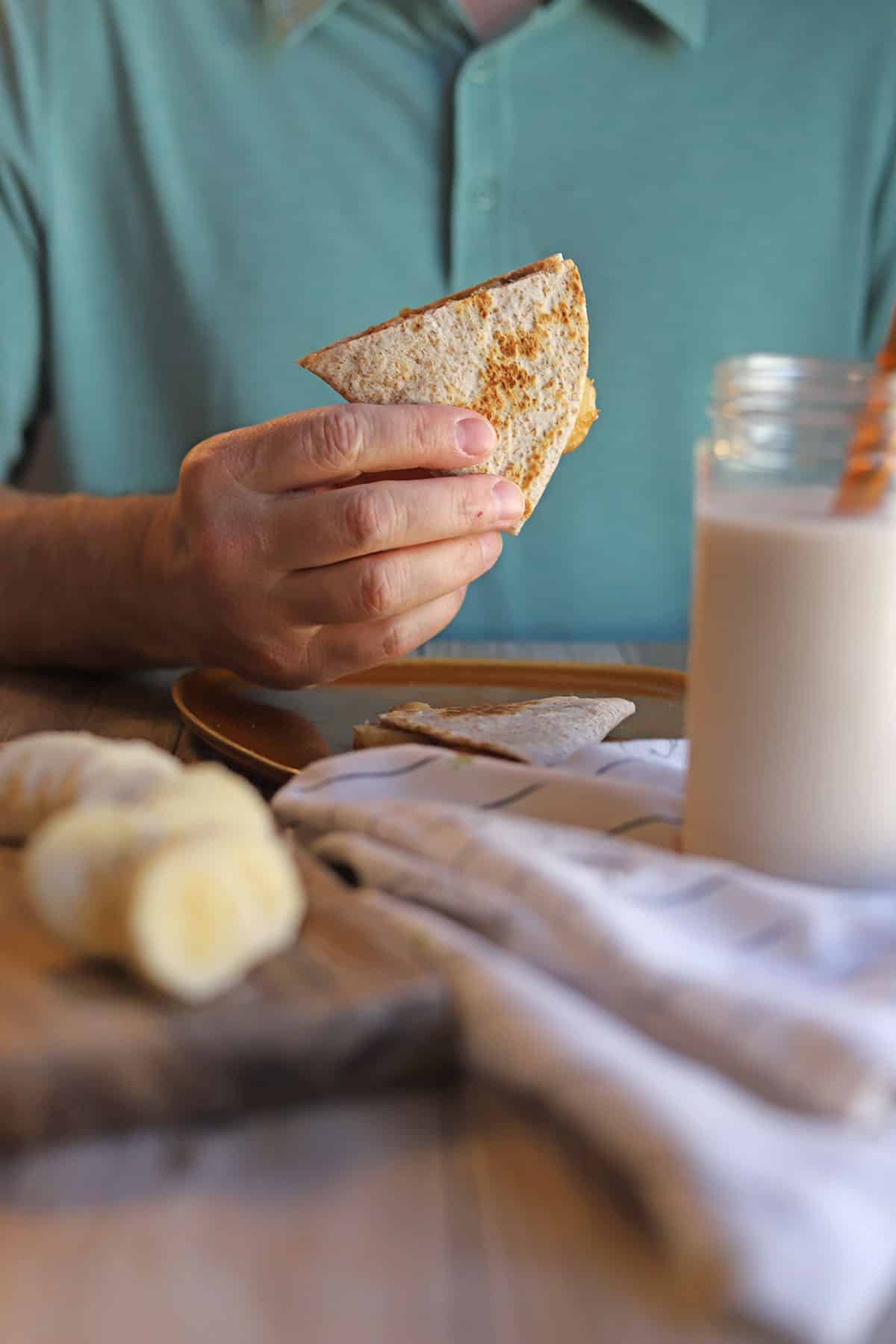 Hand holding peanut butter banana quesadilla by glass of non-dairy milk.