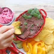 Hand dipping tortilla chip into chipotle black bean dip with cilantro garnish.