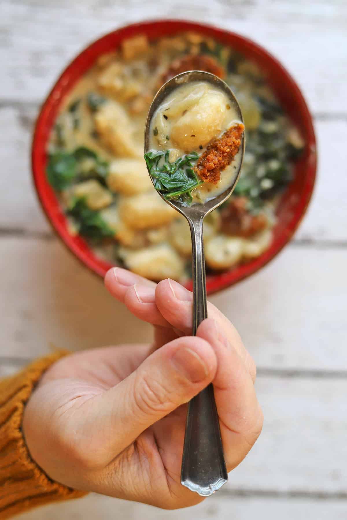Hand holding soup spoon filled with kale, sausage, and gnocchi in creamy broth.
