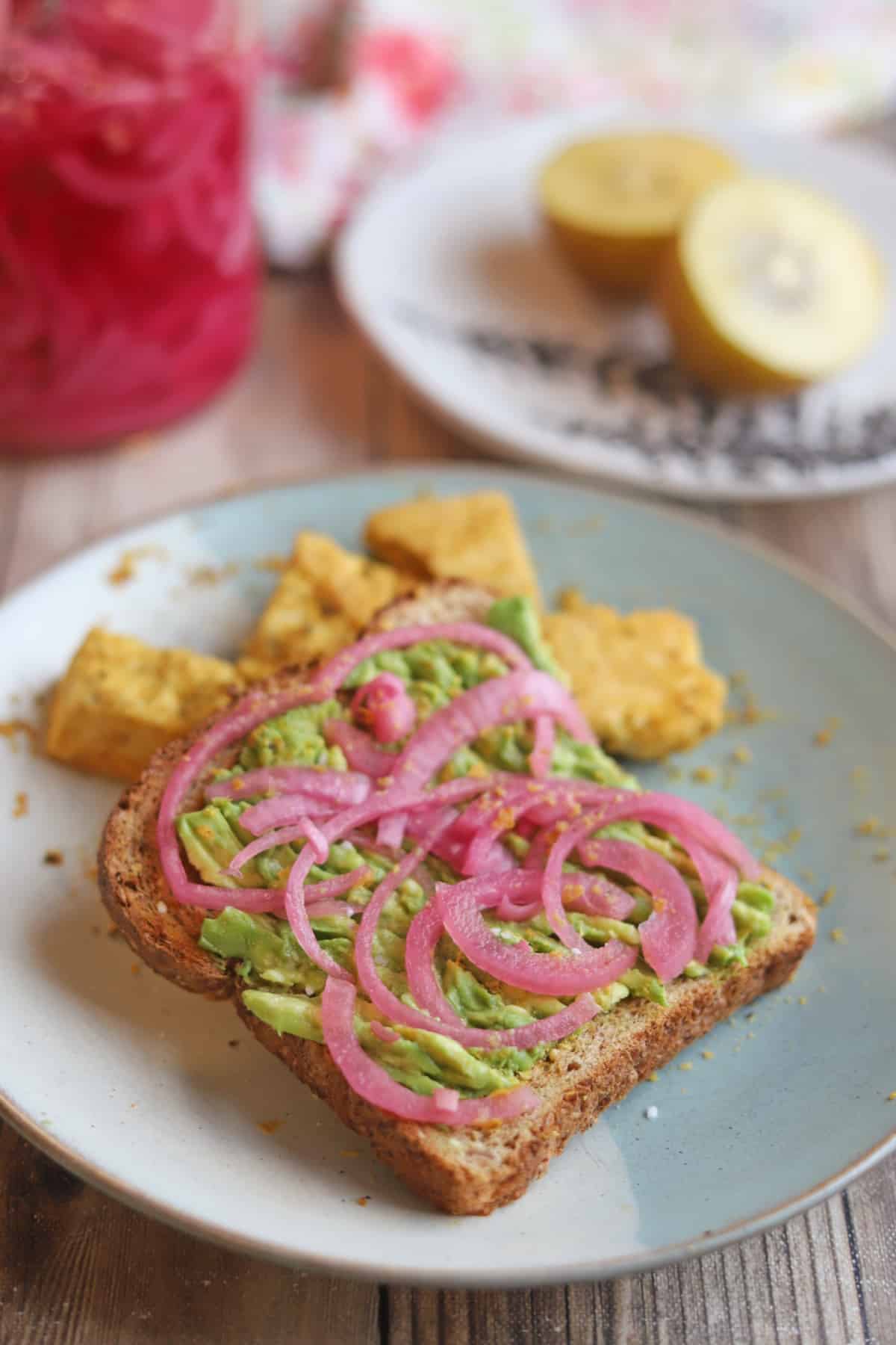 Bright pink pickled red onions on avocado toast sprinkled with nutritional yeast and salt by tofu and kiwi fruit.