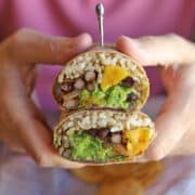 Hands holding burrito with brown rice, fried plantains, black beans, and guacamole.