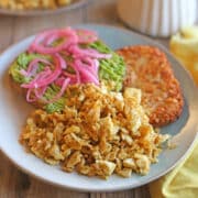 Browned silken tofu covered with nutritional yeast and black salt on plate with avocado toast and hashbrown patty.