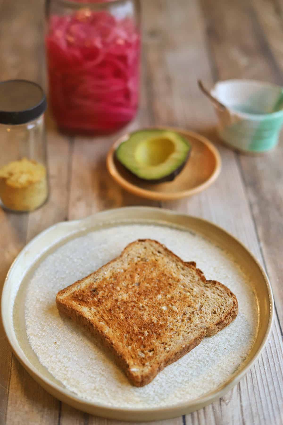 Toast on plate by avocado, nutritional yeast, pickled red onions, and salt container.