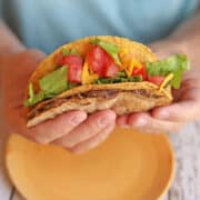 Hands holding double decker taco with refried beans, tomatoes, vegan cheddar cheese, and lettuce.