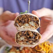 Hands holding vegan chorizo burrito sliced in half to reveal tater tots and black beans.