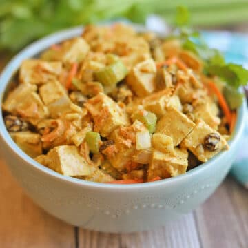 Curried tofu salad with grated carrot, celery, raisins, and vegan mayo dressing in blue bowl.