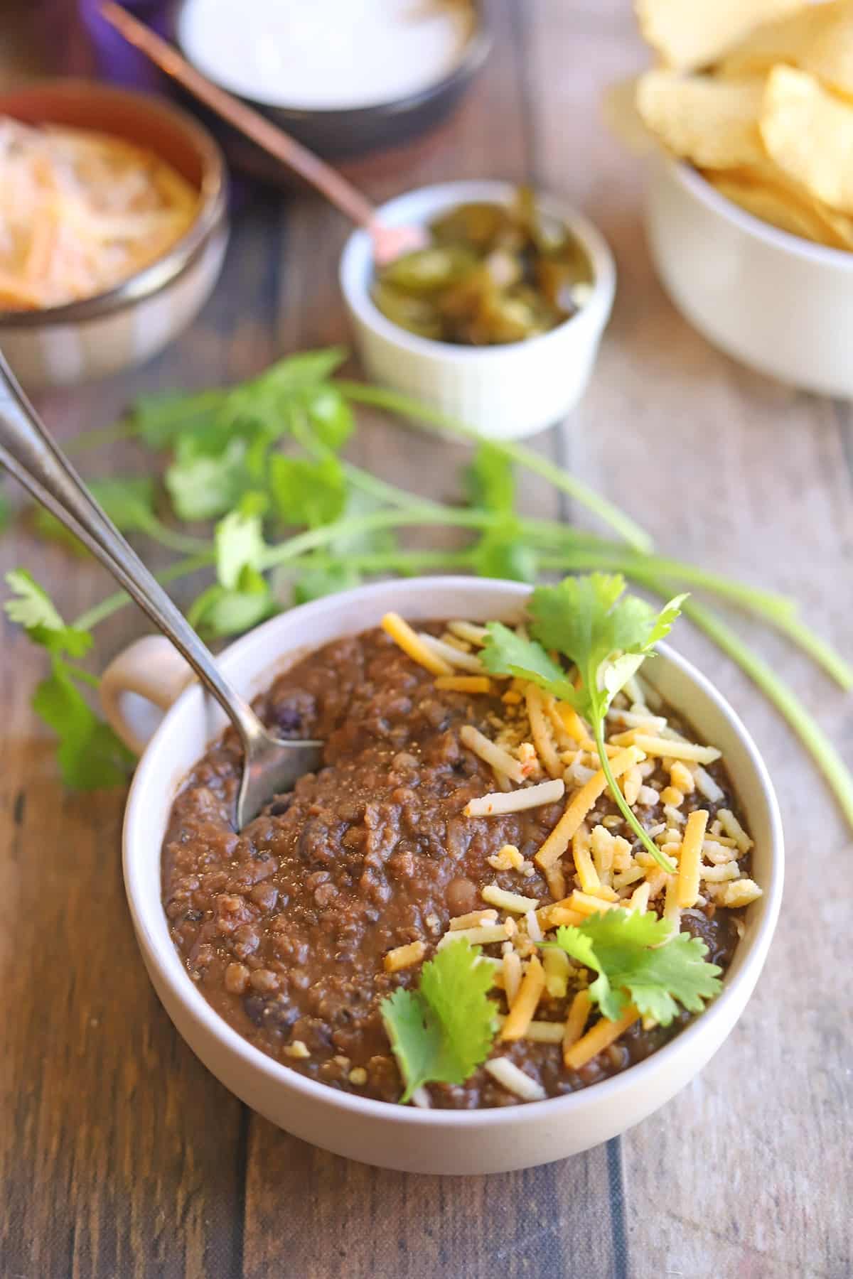 Spoon in bowl of thick lentil chili with beans, shredded non-dairy cheese, and cilantro.