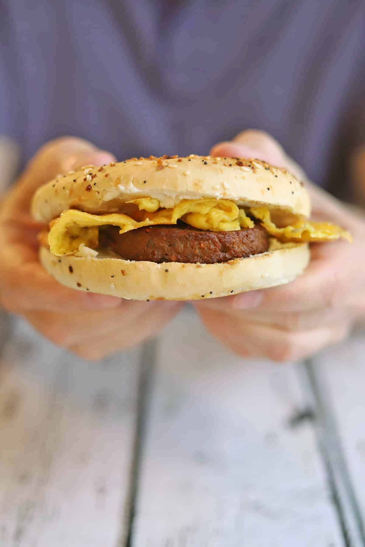 Hands holding bagel sandwich with non-dairy cream cheese, vegan sausage, and JUST Egg.