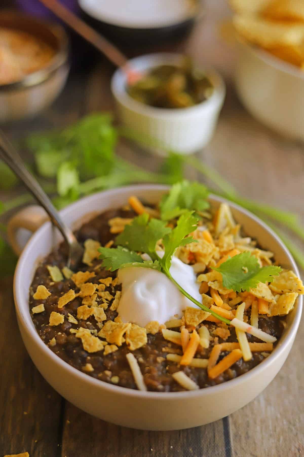 Bowl of vegan lentil chili with black beans, and topped with tortilla chips, non-dairy sour cream, and cilantro.