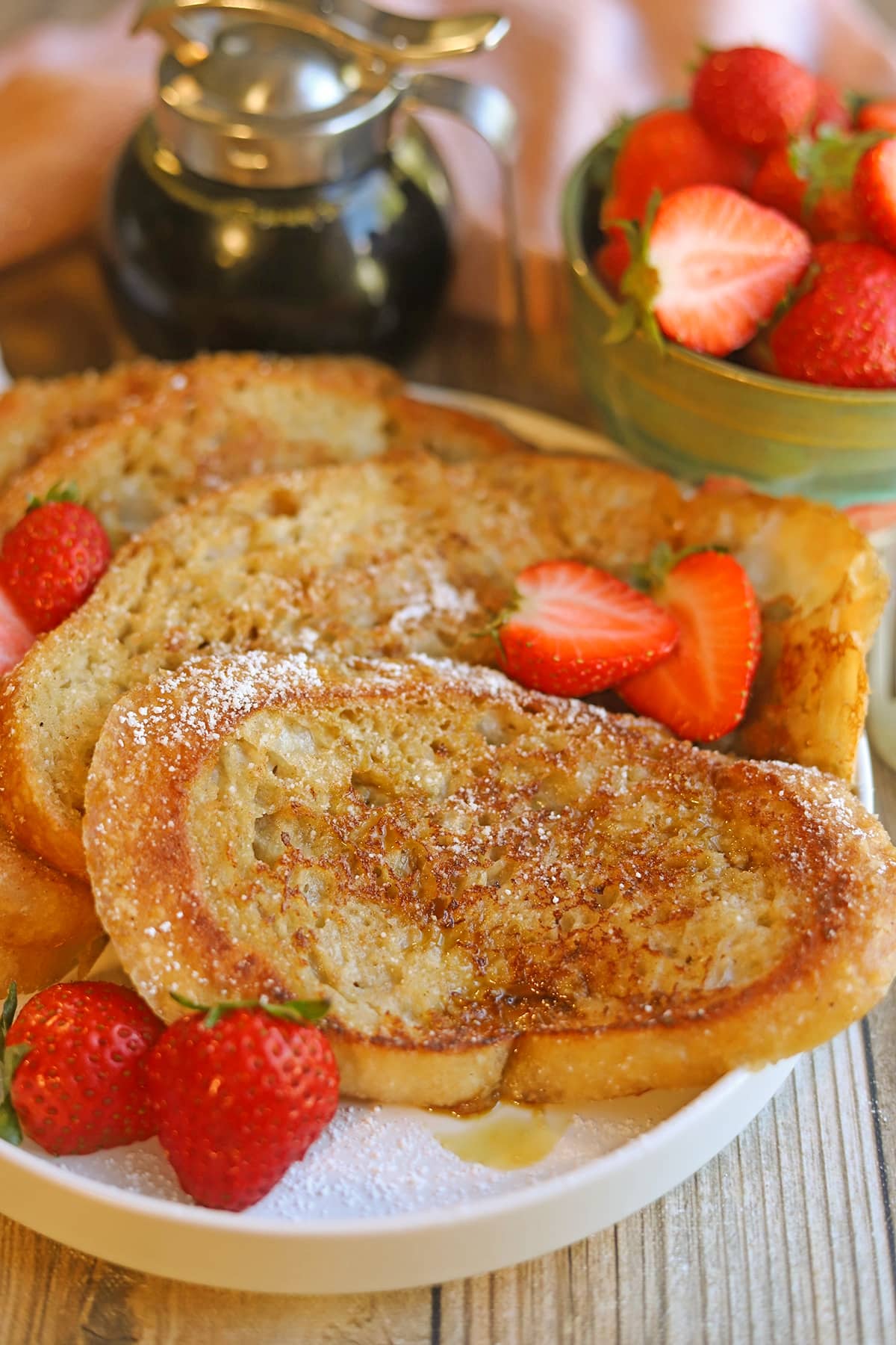 Vegan French toast on plate with strawberries, maple syrup, and powdered sugar.