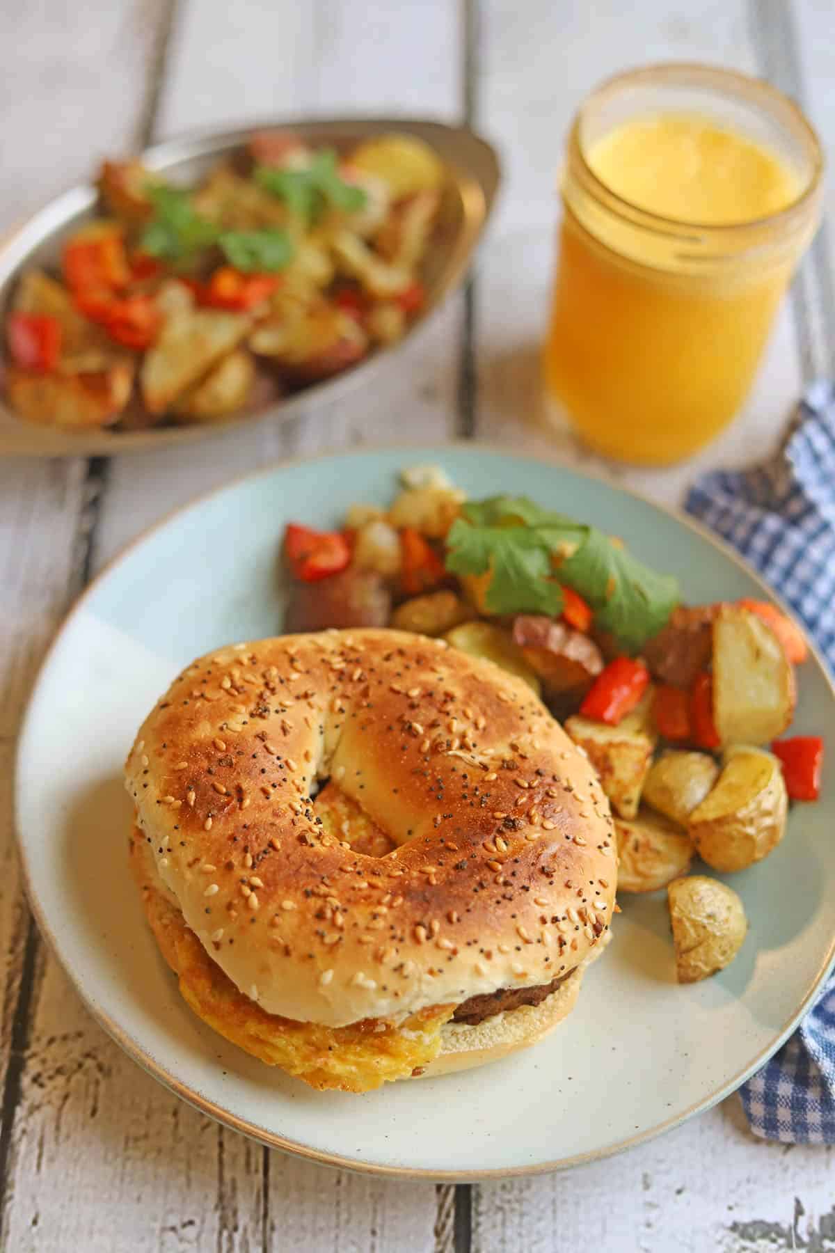 Bagel sandwich won plate with baked potato hash and glass of orange juice.