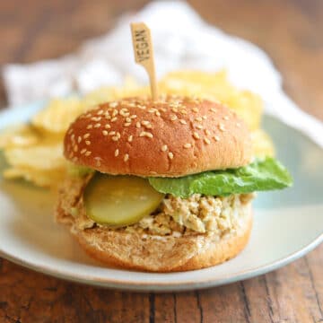 Vegan chicken salad sandwich topped with lettuce and pickles on plate.