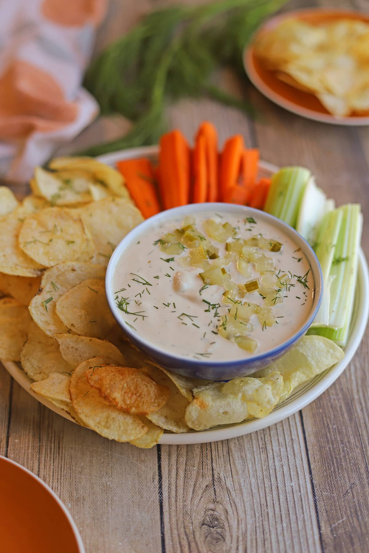 Dairy-free dill pickle dip in bowl by potato chips, carrot sticks, and celery sticks.