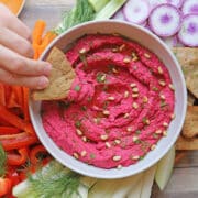 Hand dipping toasted pita chip into roasted beet hummus with toasted pine nuts and herbs.