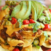 Text overlay: cadryskitchen.com, vegan nachos with cashew queso. Close-up pile of nachos with drippy dairy-free queso, soyrizo, black beans, jalapenos, and tomatoes.