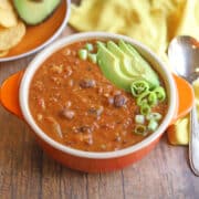 Three bean chili in orange bowl with avocado and pickled pepper garnish.