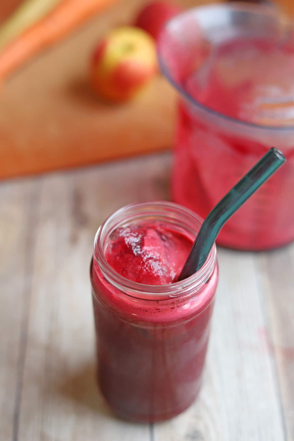 ABC juice with aqua straw on table with pitcher, apple, and carrots.
