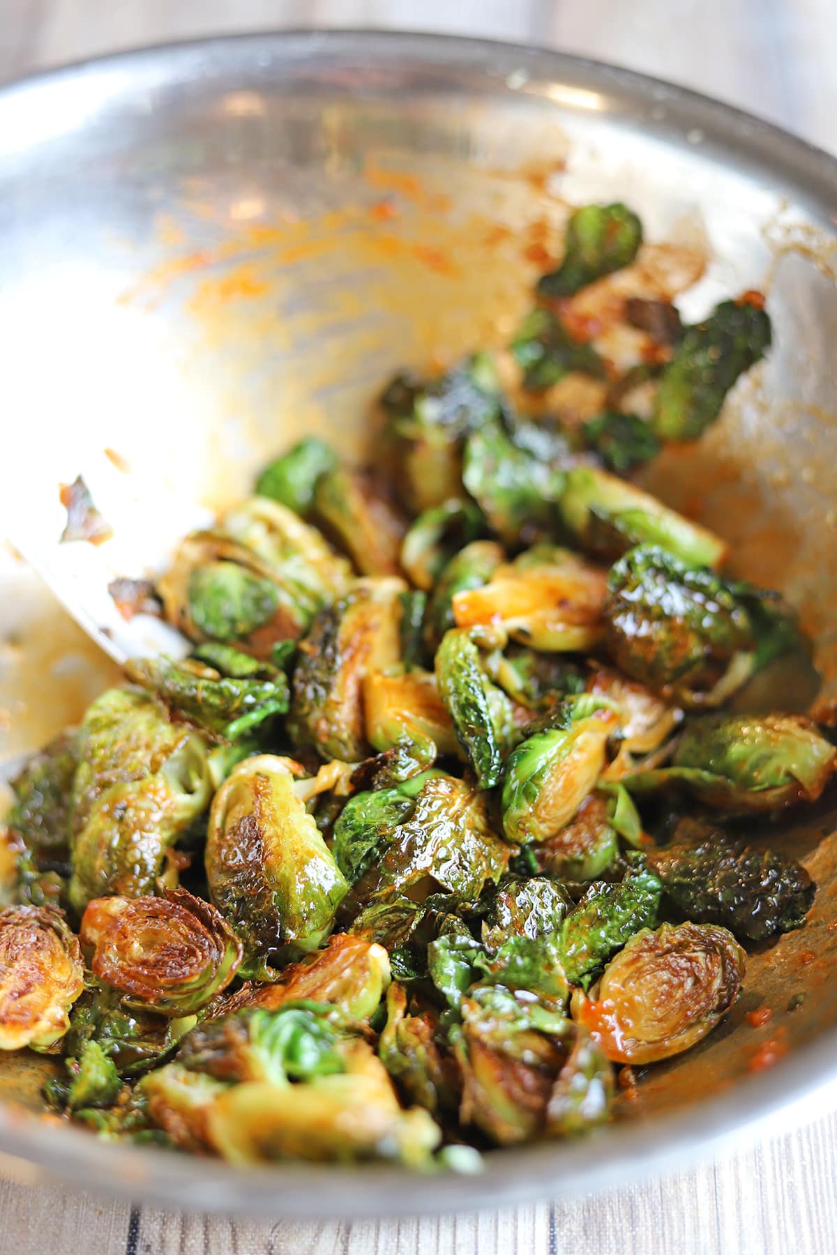Pan fried Brussels sprouts being tossed with gochujang glaze in large metal mixing bowl.