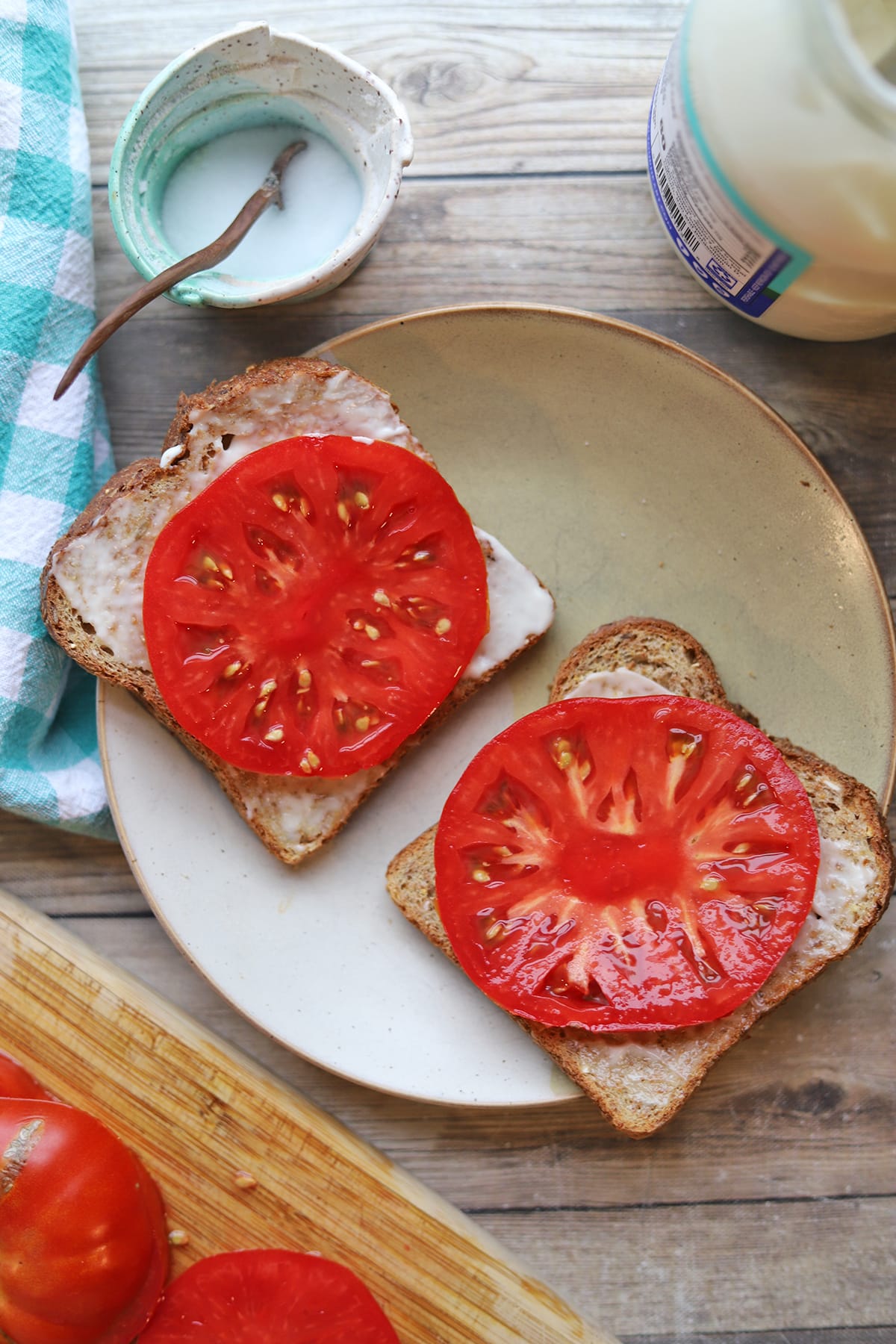 Open faced tomato sandwich on plate alongside salt, vegan mayo, and thickly sliced tomatoes.