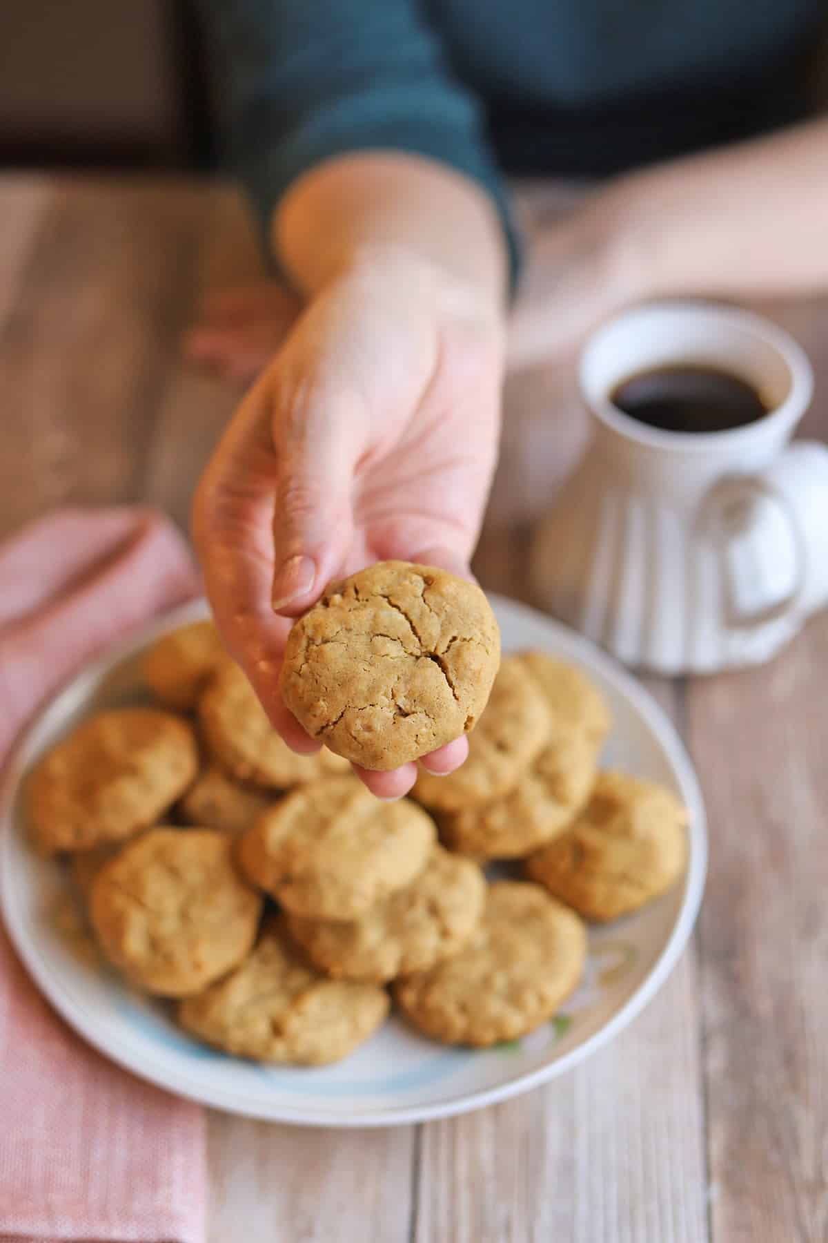 Hand holding vegan peanut butter oatmeal cookie above plate by coffee.