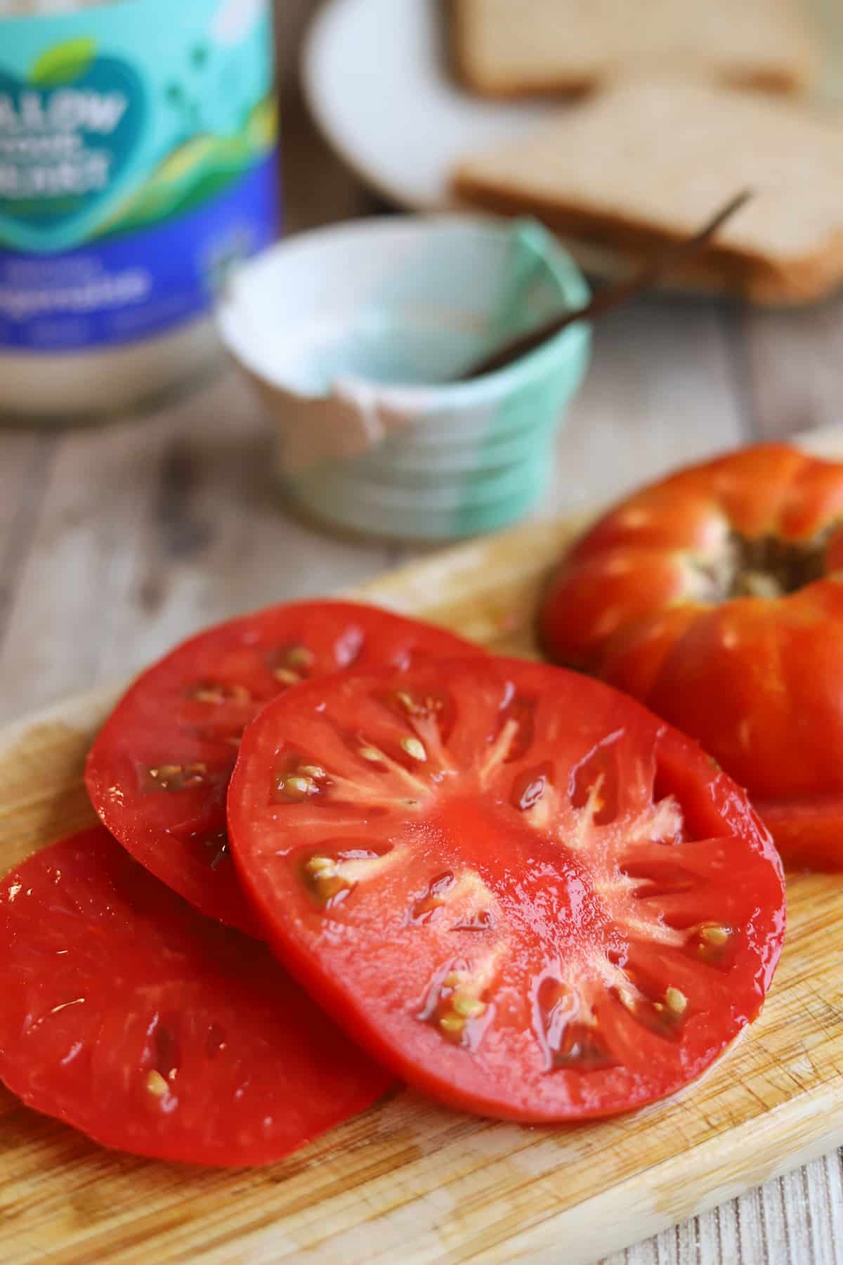 Thick slices of ripe tomato on cutting board by salt, bread, and eggless mayo.