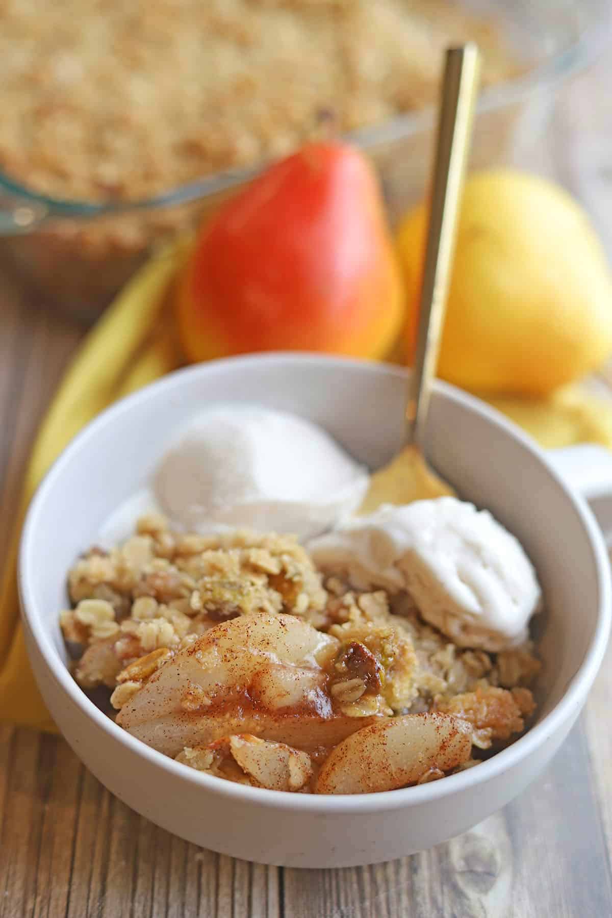 Vegan pear crisp in bowl with dollops of non-dairy ice cream. In the background, yelow and red pears.