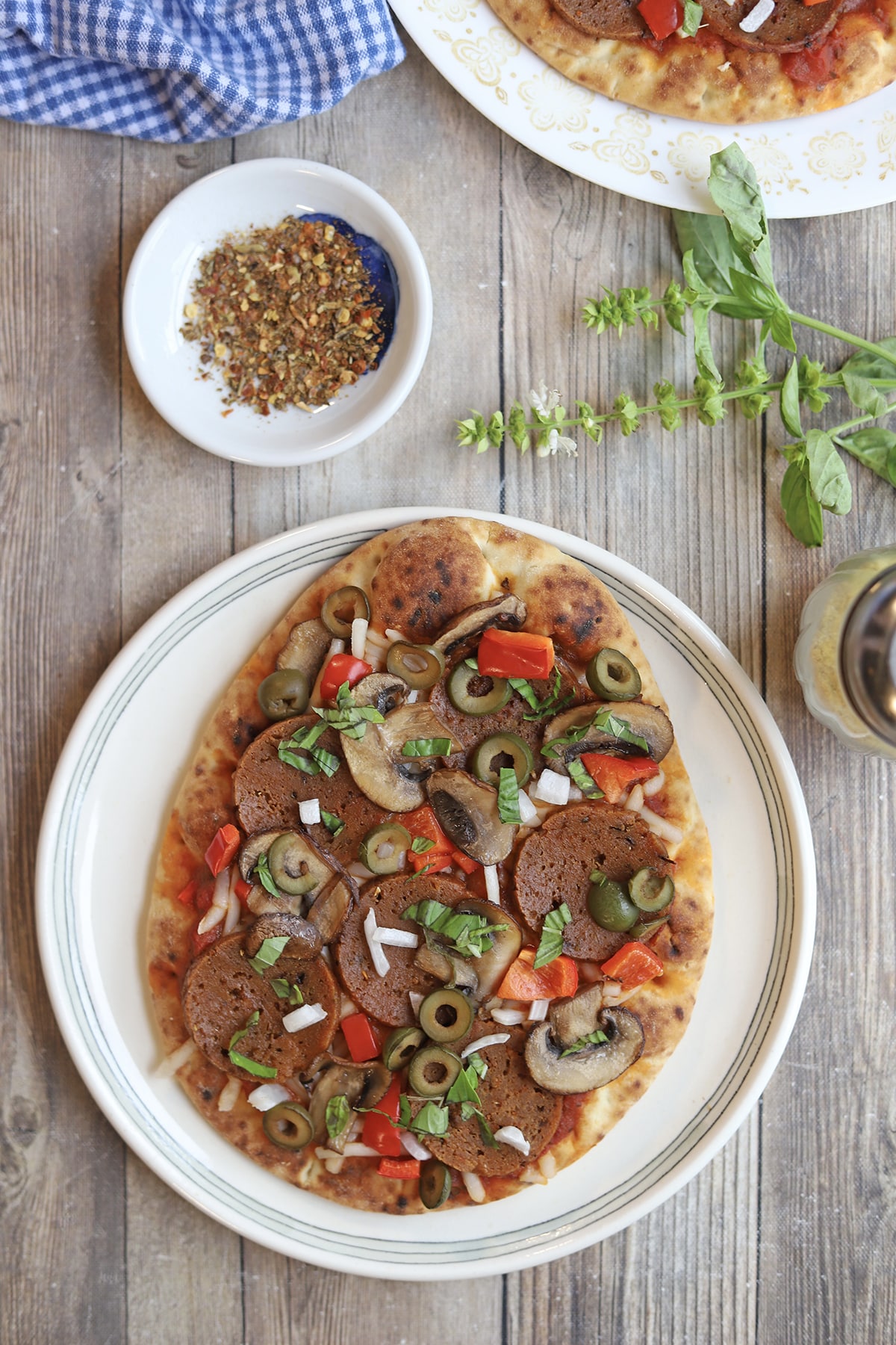 Naan flatbread pizza loaded with vegan toppings on table by red pepper flakes and fresh basil.
