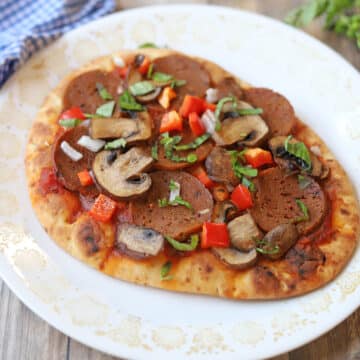 Naan flatbread pizza with mushrooms, vegan pepperoni, bell pepper, and fresh basil on white plate.