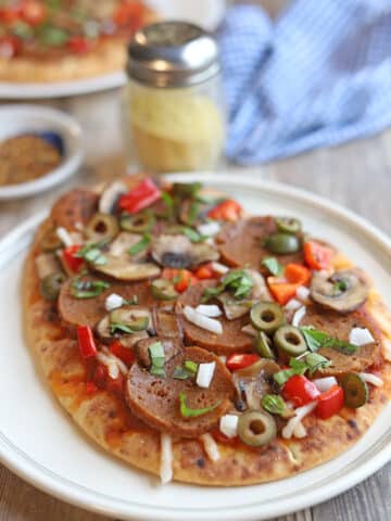 Flatbread pizza topped with vegan pepperoni, olives, red bell pepper, mushrooms, onion, and non-dairy cheese on plate by vegan parmesan cheese.