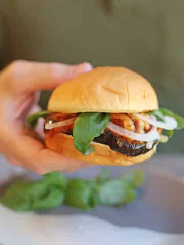 Hand holding vegan sandwich with breaded eggplant, marinara, fresh basil, and onion.