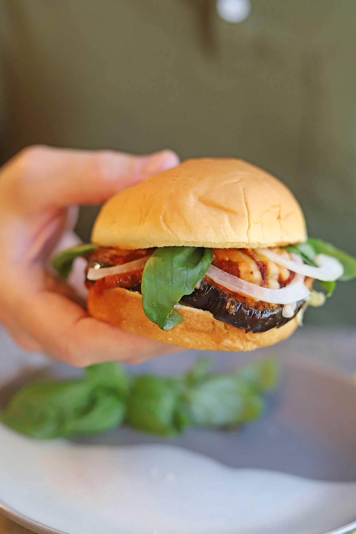 Hand holding vegan sandwich with breaded eggplant, marinara, fresh basil, and onion.