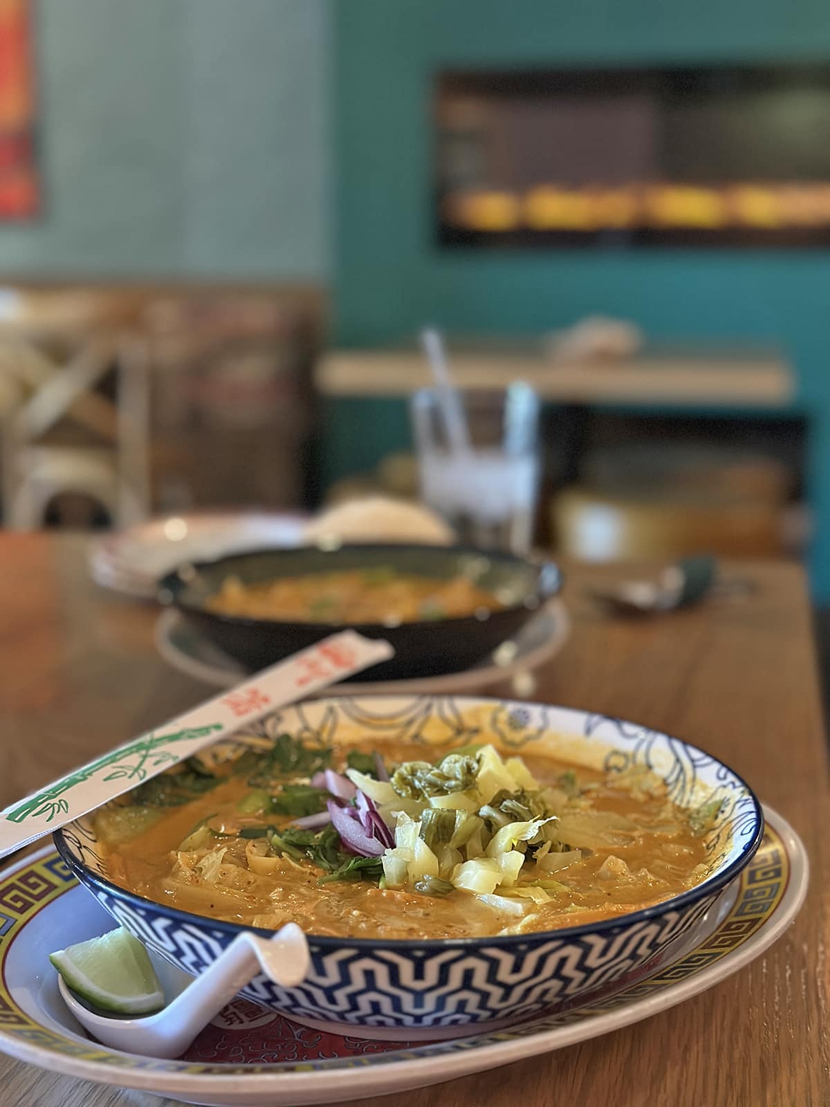 Bowl of khao soi soup with pickled vegetable garnish, soup spoon, lime wedge, and chopsticks on table at Miss Phay in Davenport, Iowa.