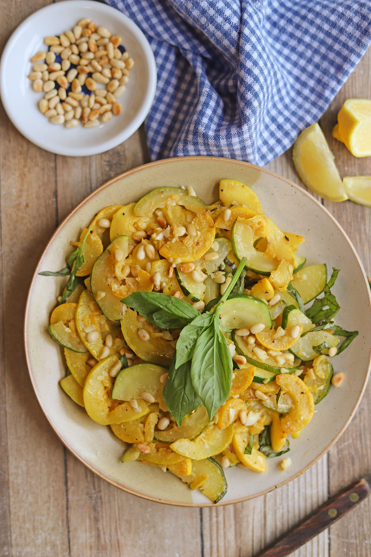Recipe with summer squash and zucchini on serving plate by lemons and toasted pine nuts.