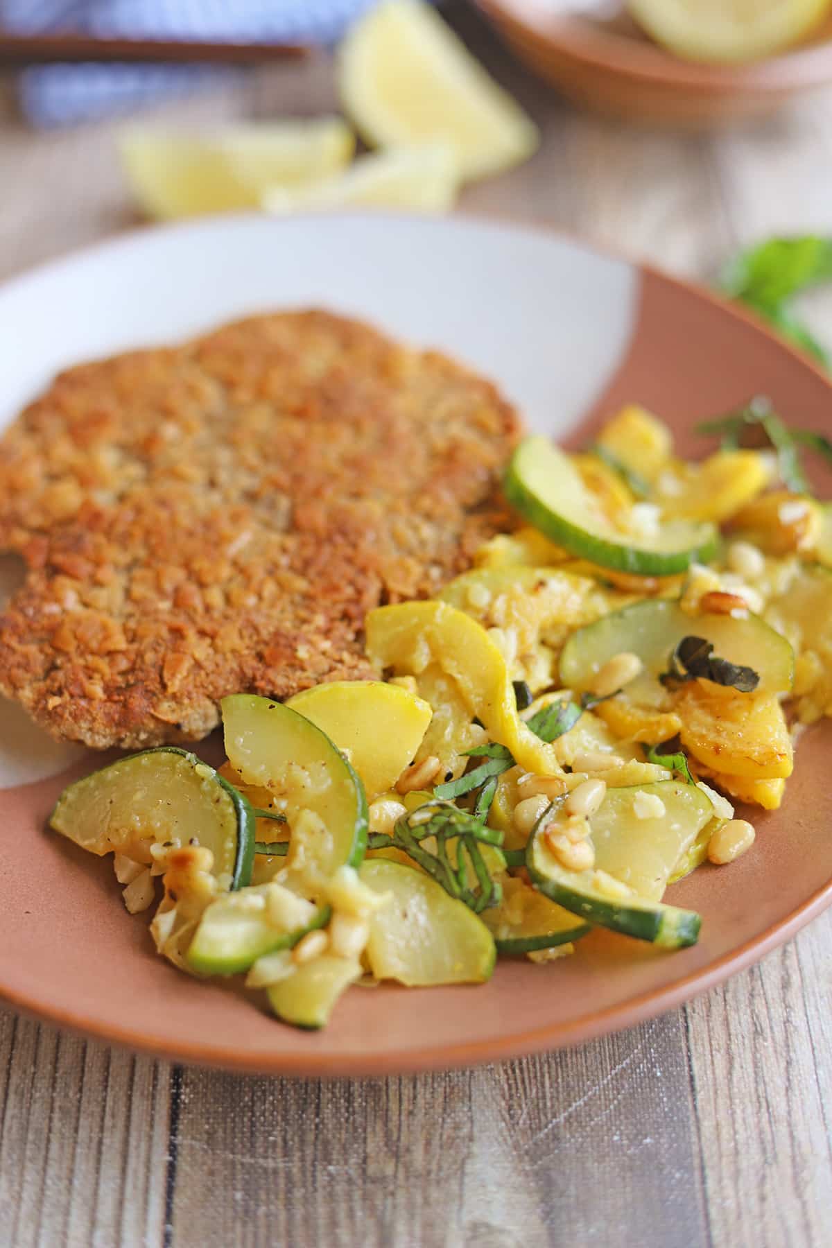 Yellow summer squash and zucchini garnished with basil and toasted pine nuts on plate with vegan schnitzel.
