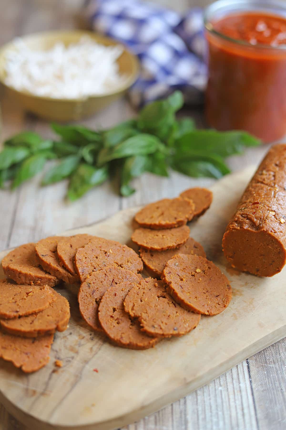 Sliced log of vegan pepperoni on cutting board by marinara, fresh basil, and non-dairy cheese.