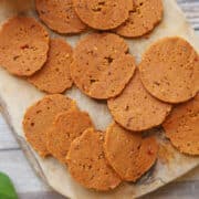 Slices of seitan pepperoni dotted with fennel seeds and mustard seeds on cutting board.