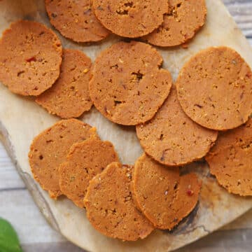 Slices of seitan pepperoni dotted with fennel seeds and mustard seeds on cutting board.