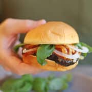 Hand holding vegan eggplant parmesan sandwich on puffy bun with fresh basil, onion, marinara, and non-dairy cheese.