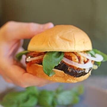 Hand holding vegan eggplant parmesan sandwich on puffy bun with fresh basil, onion, marinara, and non-dairy cheese.