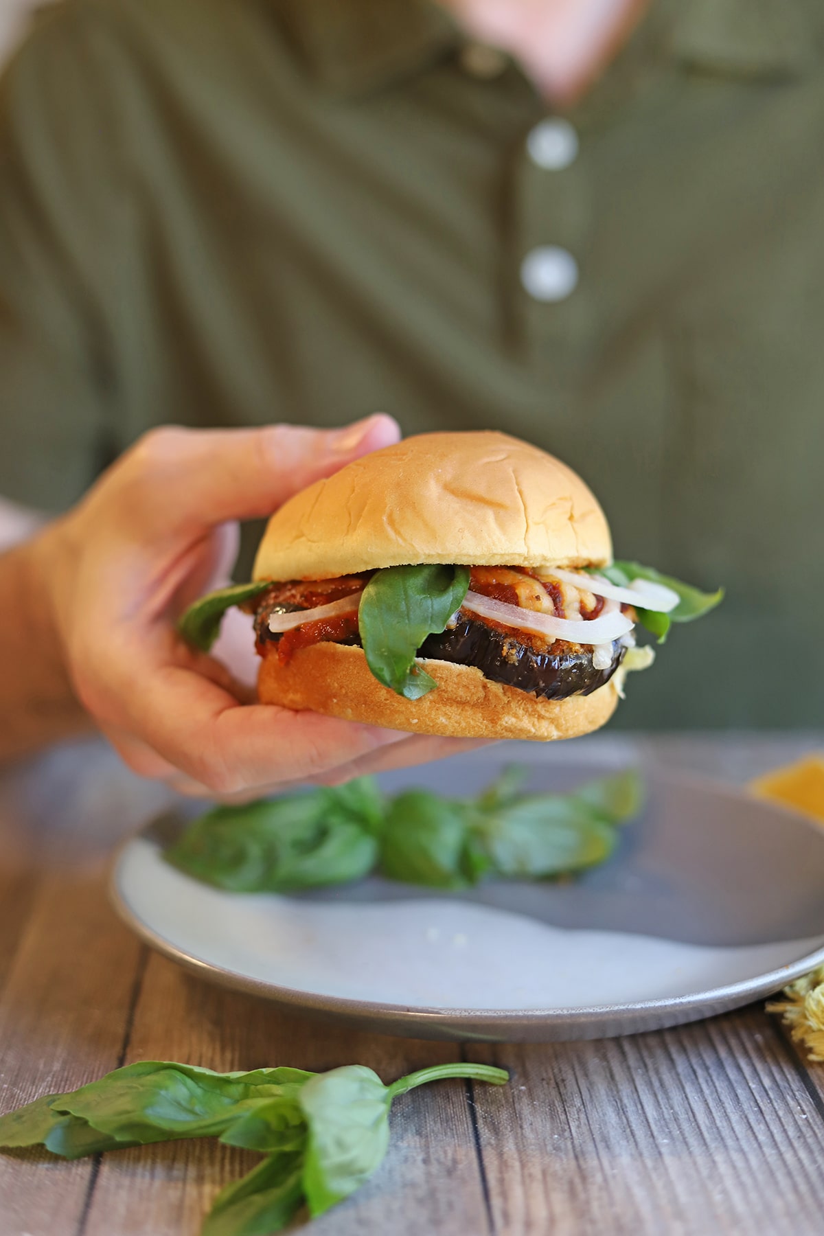Hand holding vegan eggplant parmesan sandwich with fresh basil and onion.