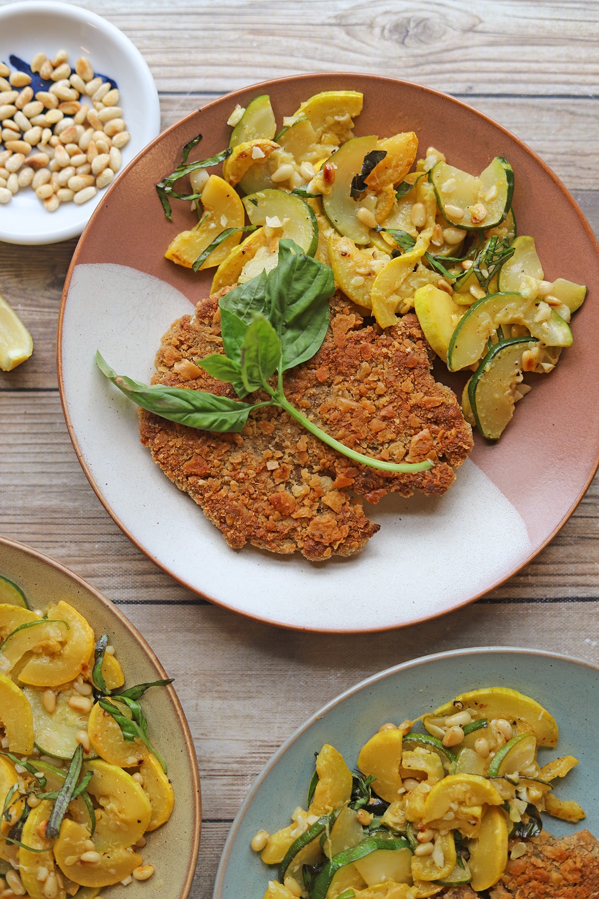 Vegan schnitzel on plate with sauteed zucchini and summer squash topped with basil and toasted pine nuts.