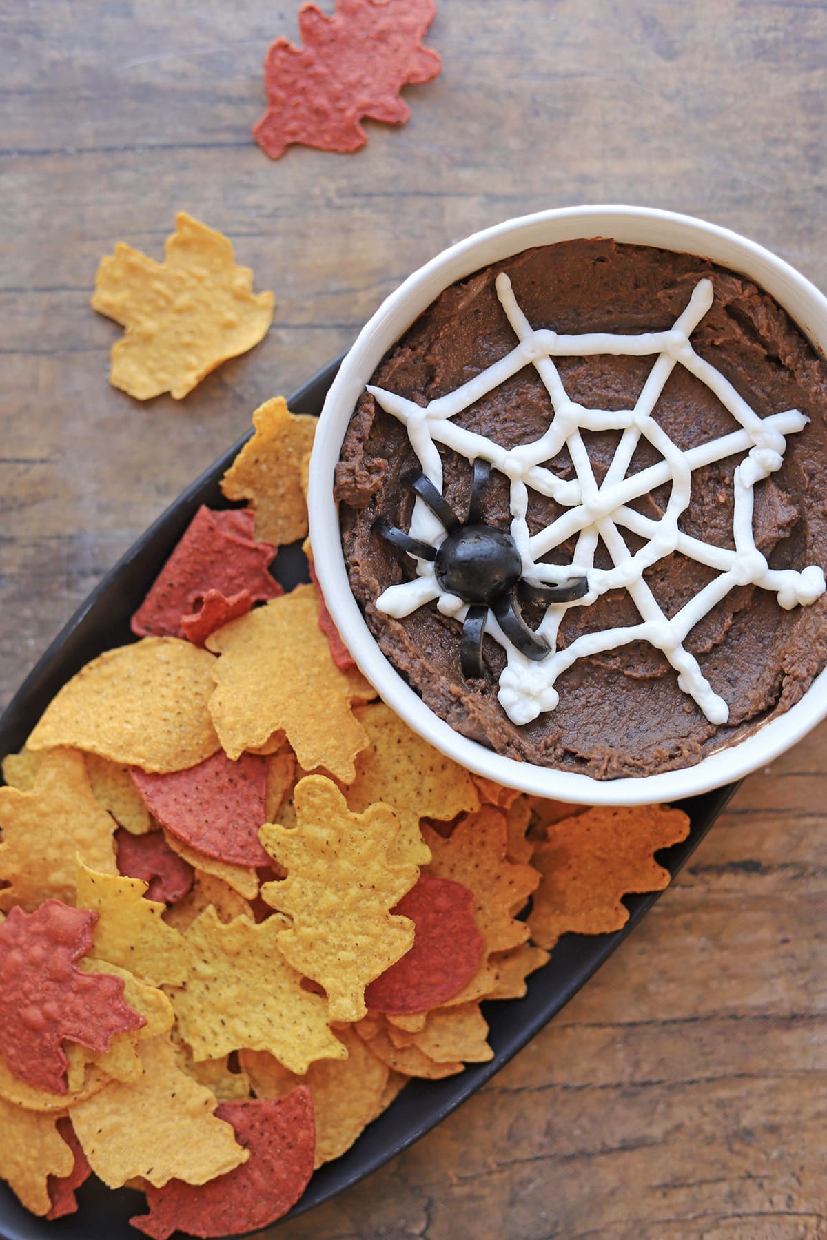 Leaf-shaped tortilla chips on platter with chipotle black bean dip, sour cream web, and olive spider.