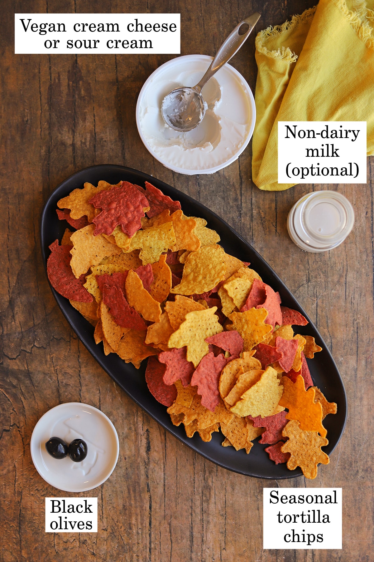 Labeled ingredients for Halloween dip toppings with vegan cream cheese, non-dairy milk, seasonal tortilla chips, and black olives.