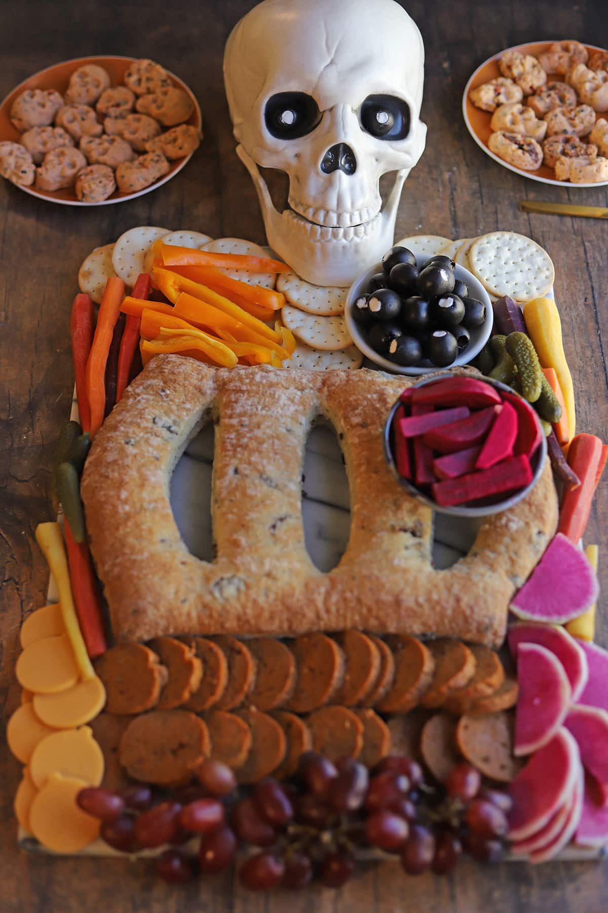 Vegan charcuterie for Halloween. Skeleton shaped snacking board with vegan butter skulls, cheddar pumpkins, pepperoni intestines, turnip heart, black olive eyeballs, and cracker shoulder blades.