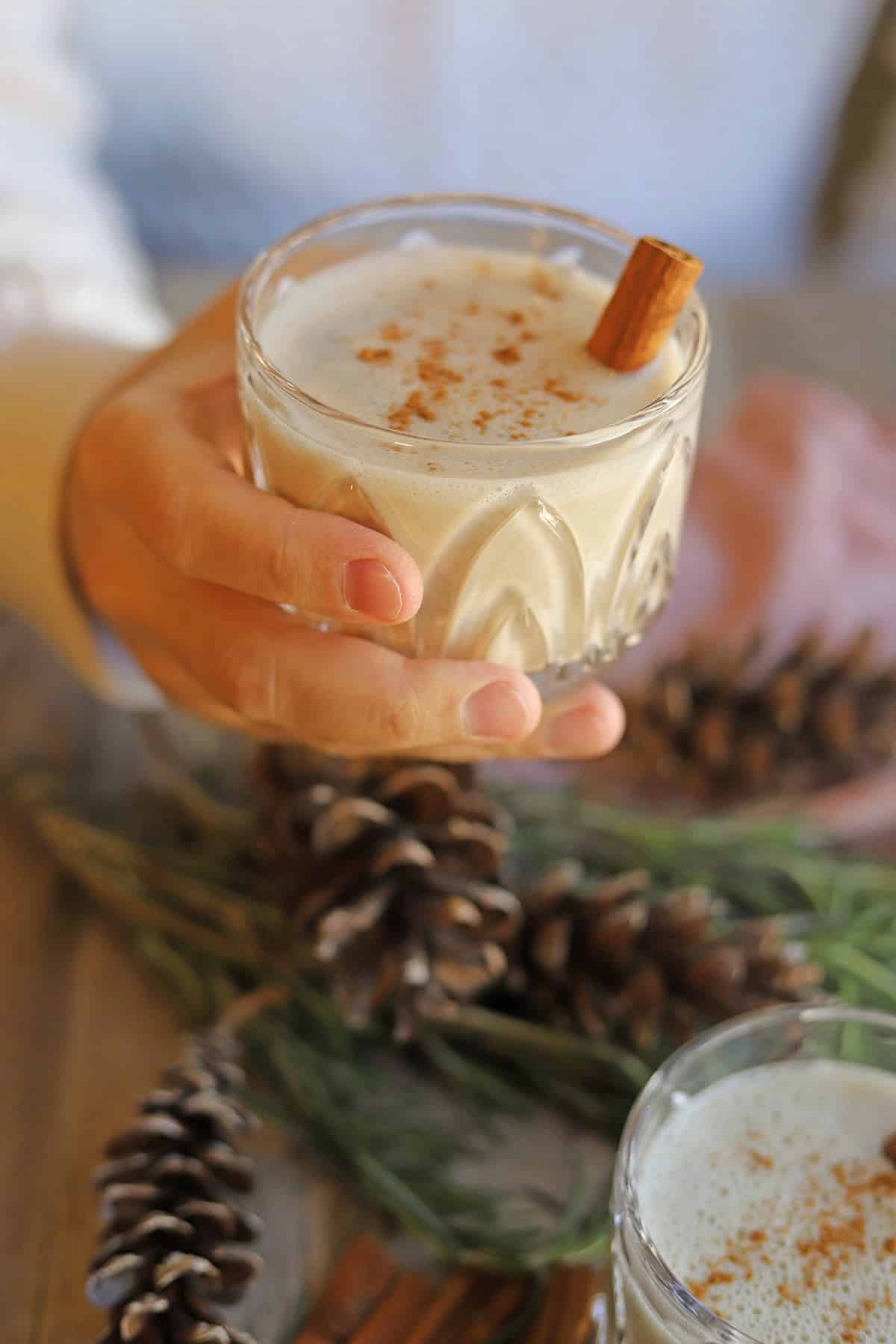 Hand holding glass of cashew milk nog with cinnamon over pinecones and greenery.