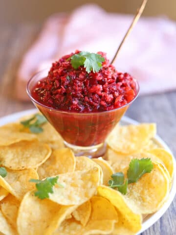Glass cocktail glass with cranberry salsa garnished with cilantro on plate of tortilla chips.