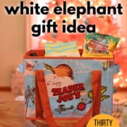 Text overlay: Trader Joe's white elephant gift idea, thirty dollar limit, cadryskitchen.com. Trader Joe's bag filled with snacks and treats in front of pink Christmas tree.