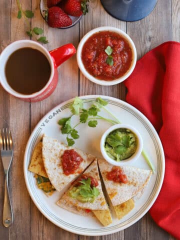 Vegan breakfast quesadilla on plate with guacamole and salsa, alongside coffee and strawberries.